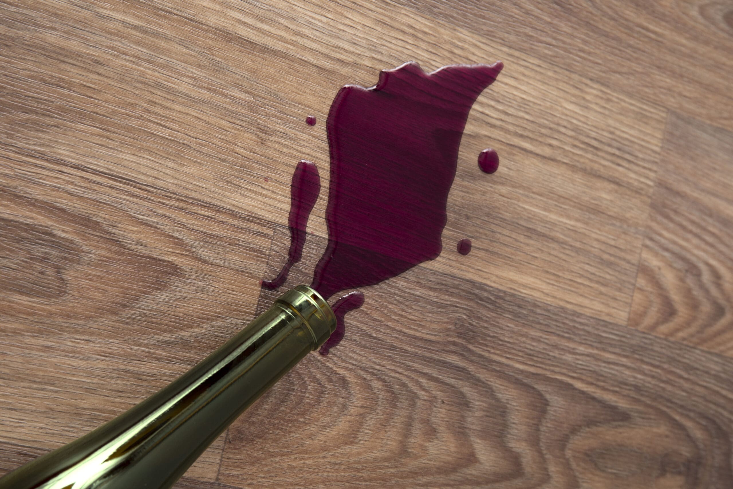 Rotwein läuft aus einer Flasche auf Parkettboden – frischer Rotweinfleck auf Holz, der ohne richtige Behandlung tief in das Parkett eindringen kann.